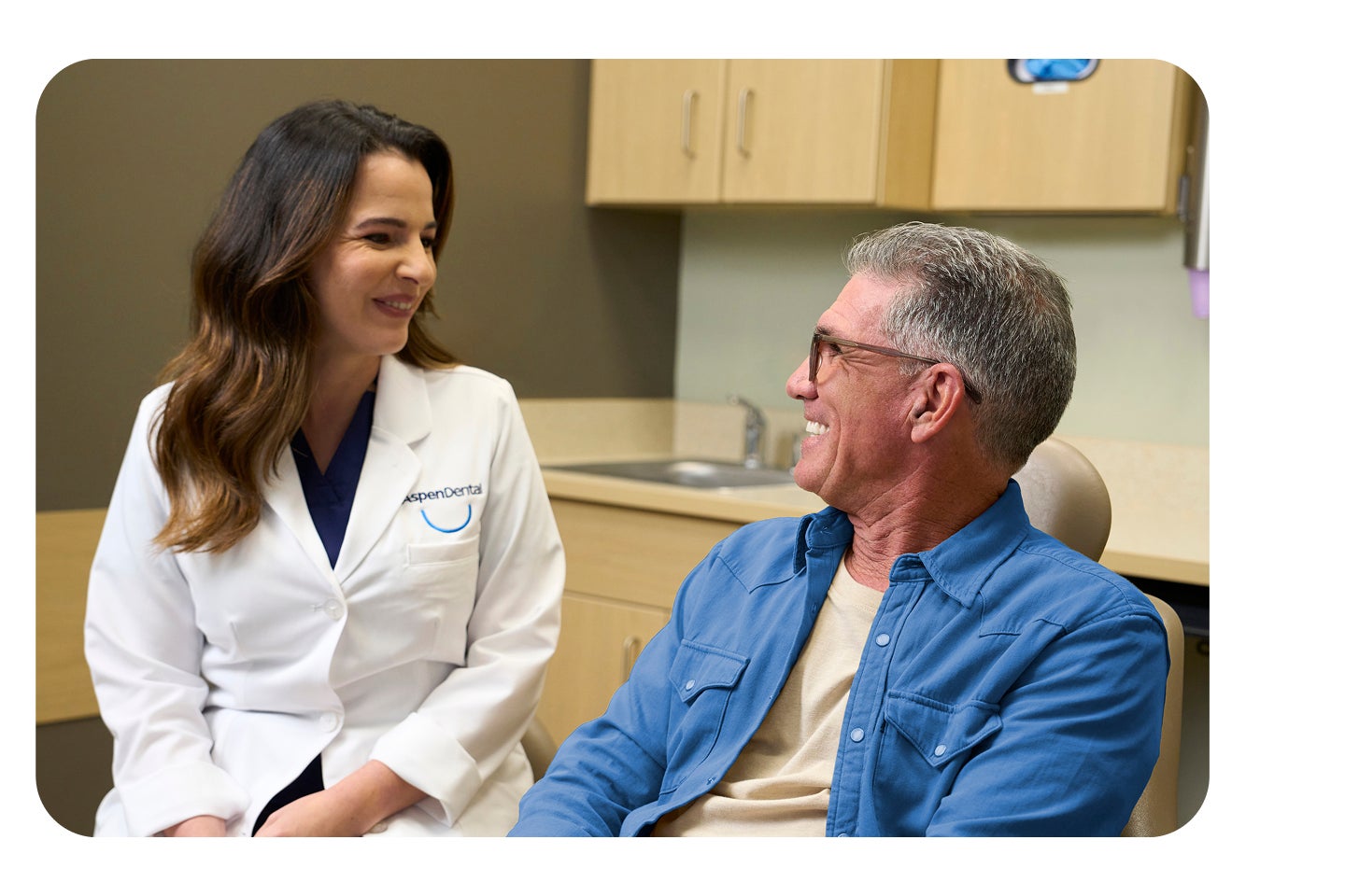 An Aspen Dental dentist in a white coat smiles while speaking with a male patient seated in a dental exam chair during a single-tooth implant consultation.