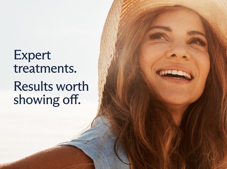 A smiling woman holding onto a sunhat on a sunny beach, overlaid with text which reads "Expert treatments. Results worth showing off."
