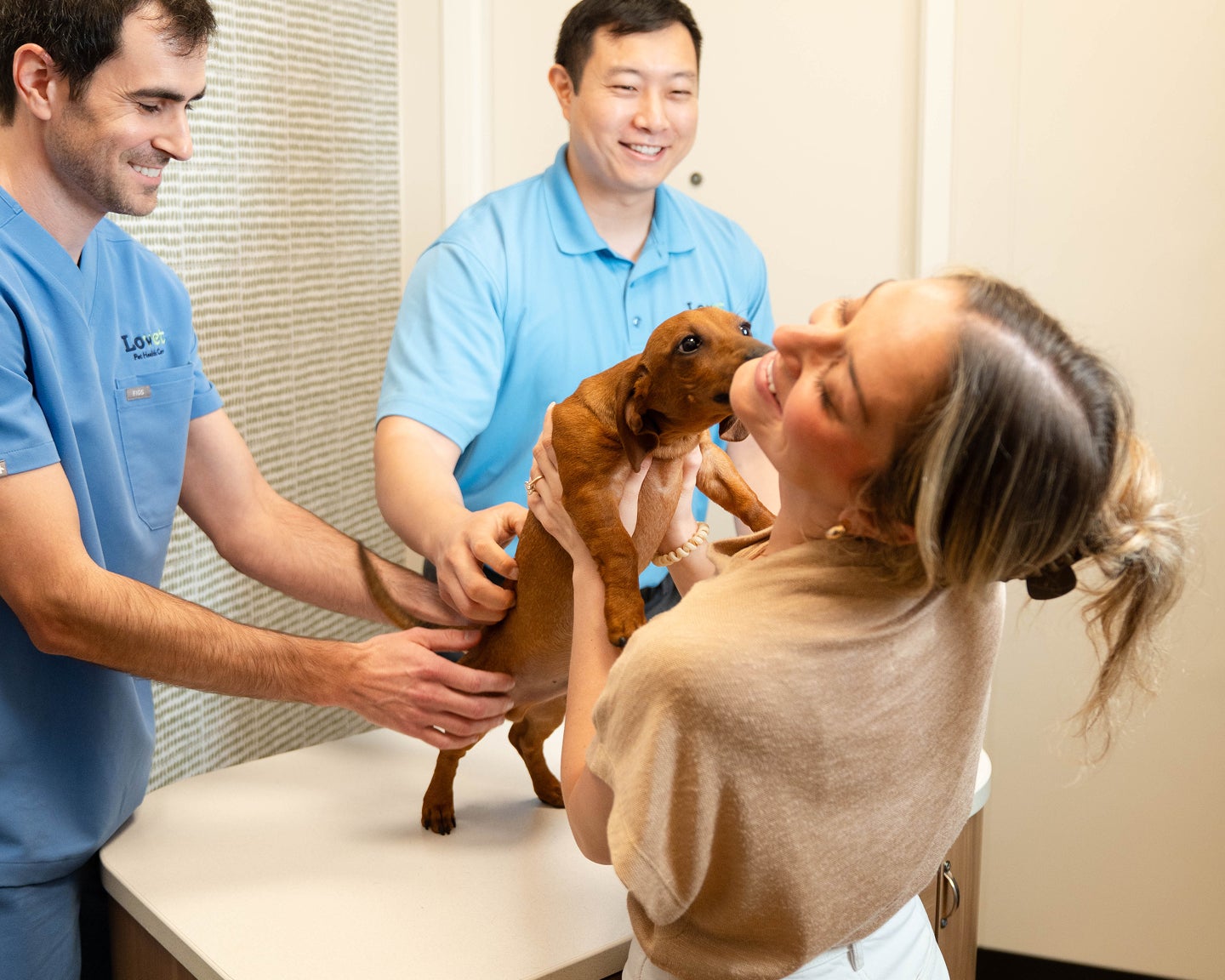 A Lovet veterinarian and team member help examine a small brown dog while its pet parent smiles and cuddles the dog during a visit at a Lovet hospital. The moment highlights a warm, reassuring vet care experience and the bond between pets, pet parents, and the care team.