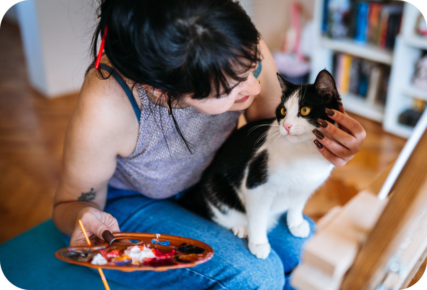 A woman sitting on the floor holding a black and white cat while painting on a small palette.