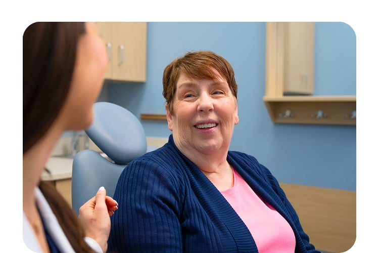 A smiling female patient wearing a pink shirt and navy cardigan talks with her Aspen Dental dentist during a friendly consultation in a dental exam room.