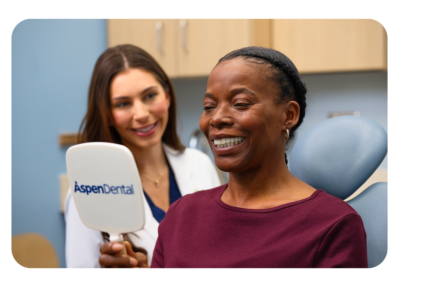 A smiling female patient looks into an Aspen Dental hand mirror while a dentist in a white coat stands beside her during a cosmetic dentistry visit.