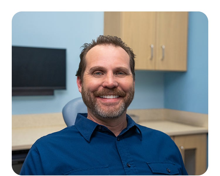 A smiling male patient sits in a dental exam chair wearing a blue shirt, with cabinetry and dental equipment visible in the background of the Aspen Dental office.