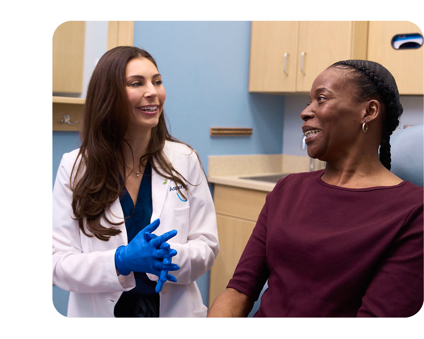 An Aspen Dental doctor in a white coat and blue gloves is smiling while having a discussion with a woman sitting in the dental chair.