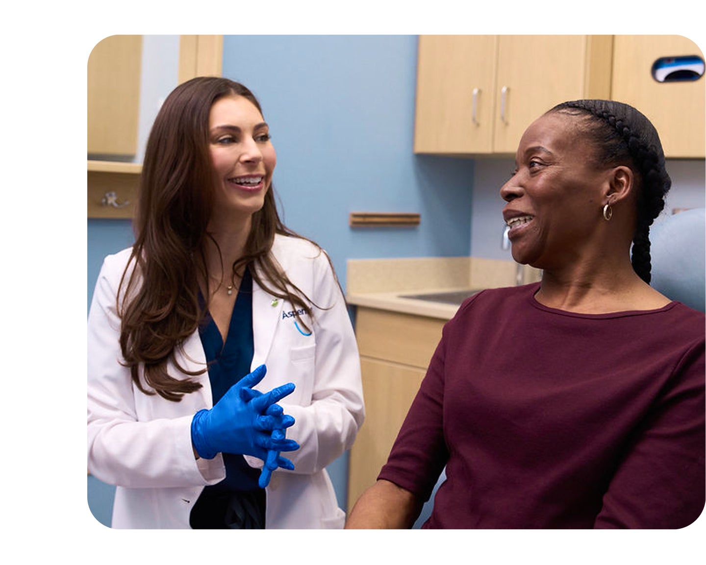  An Aspen Dental doctor in a white coat and blue gloves is smiling while having a discussion with a woman sitting in the dental chair.