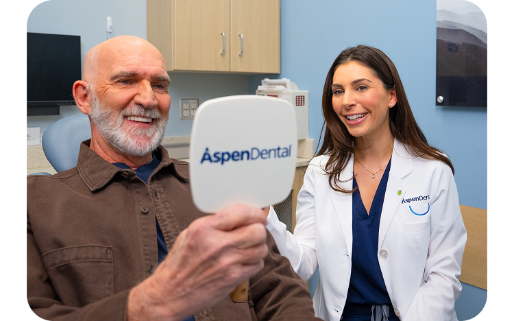 A smiling male patient looks into an Aspen Dental mirror while a dentist in a white coat smiles beside him during a dental appointment.