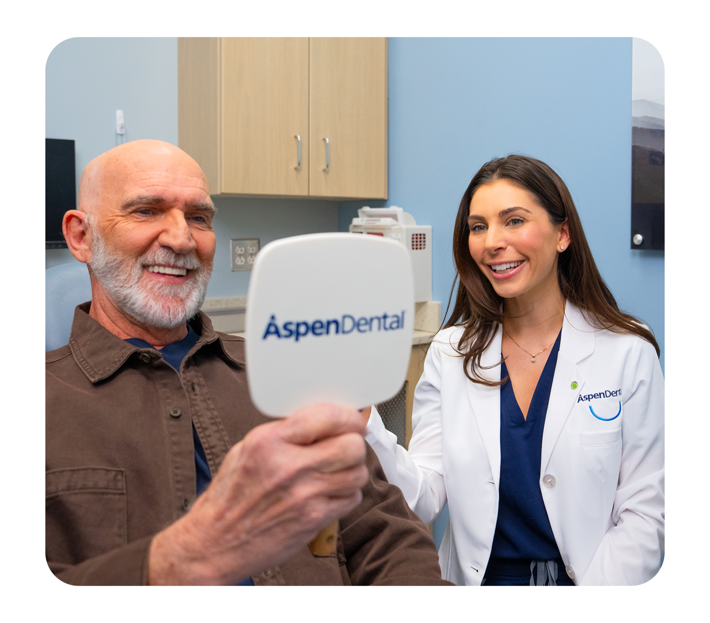 A smiling male patient looks into an Aspen Dental mirror while a dentist in a white coat smiles beside him during a dental appointment.