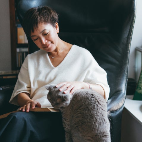 A pet parent in a leather armchair reads a book and pets a gray cat.