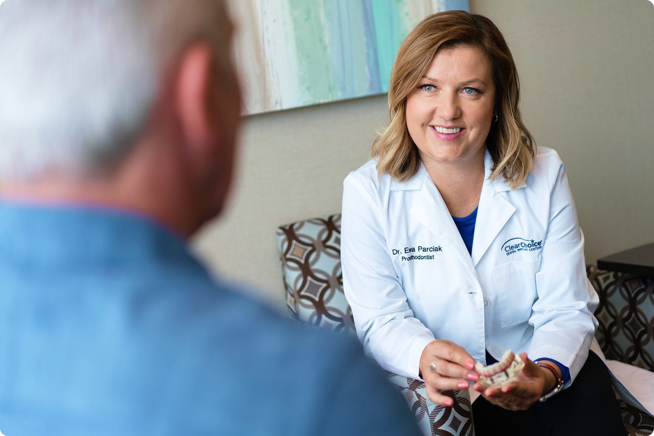 A ClearChoice doctor explains treatment options to a patient during a consultation, using a dental model to guide the conversation about insurance coverage, costs, and implant procedures.