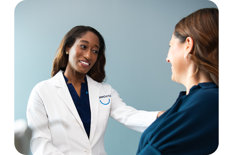 An Aspen Dental dentist in a white coat gently speaks with a seated patient, offering reassurance during a consultation in a modern exam room.
