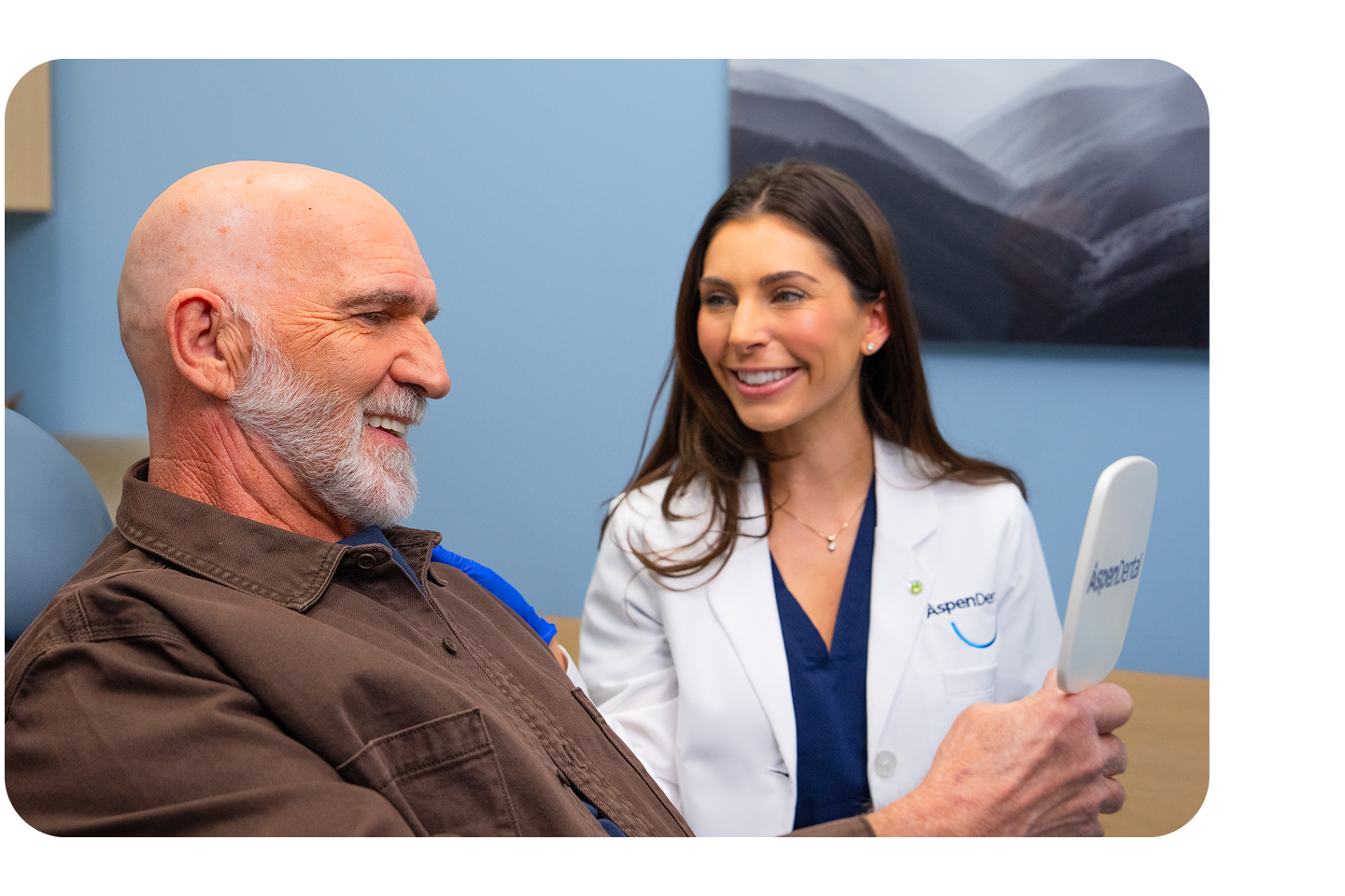 Elderly male patient smiling while holding a mirror as an Aspen Dental dentist smiles beside him during a consultation