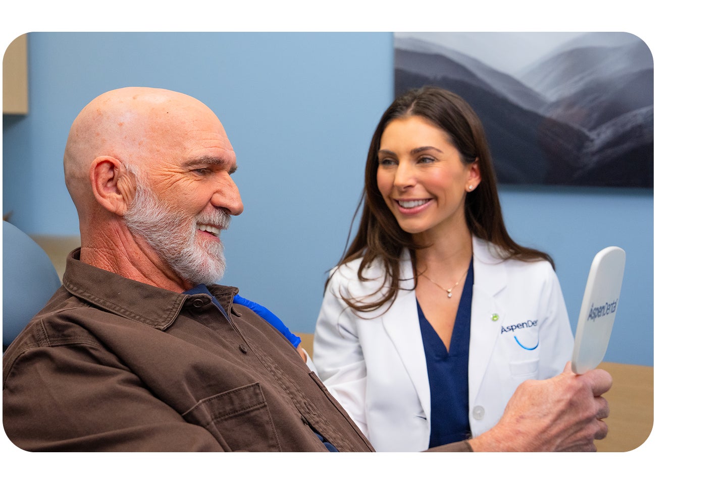 Elderly male patient smiling while holding a mirror as an Aspen Dental dentist smiles beside him during a consultation