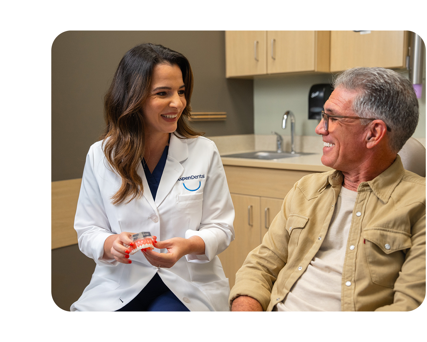 A dental professional in a white coat holds a dental model while sitting next to an older man in a dental office.