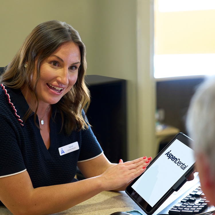 An Aspen Dental team member smiles while reviewing information on a tablet with a patient across the desk during a consultation.