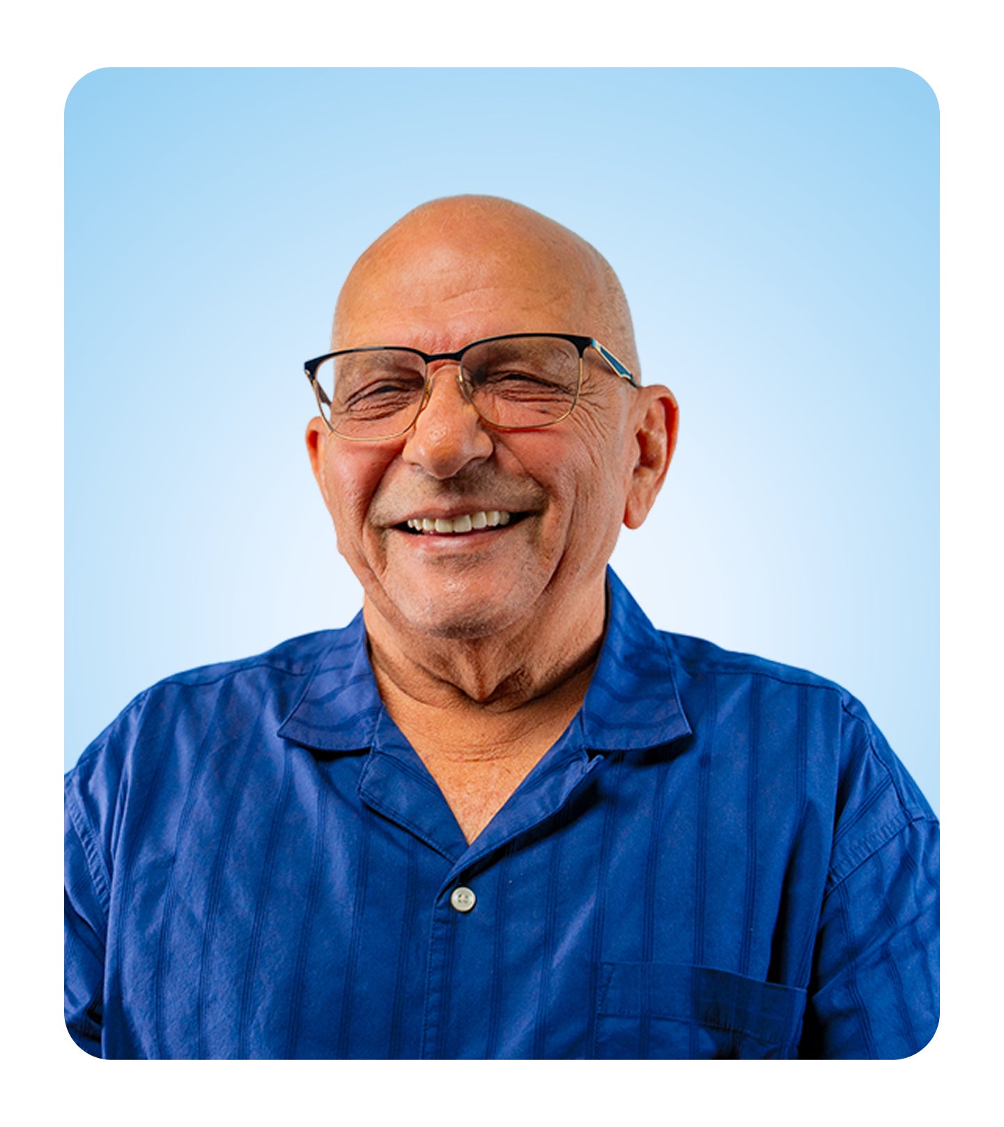 A close-up shot of a smiling senior man with glasses and a shaved head, wearing a blue shirt. He is laughing and looking directly at the camera.