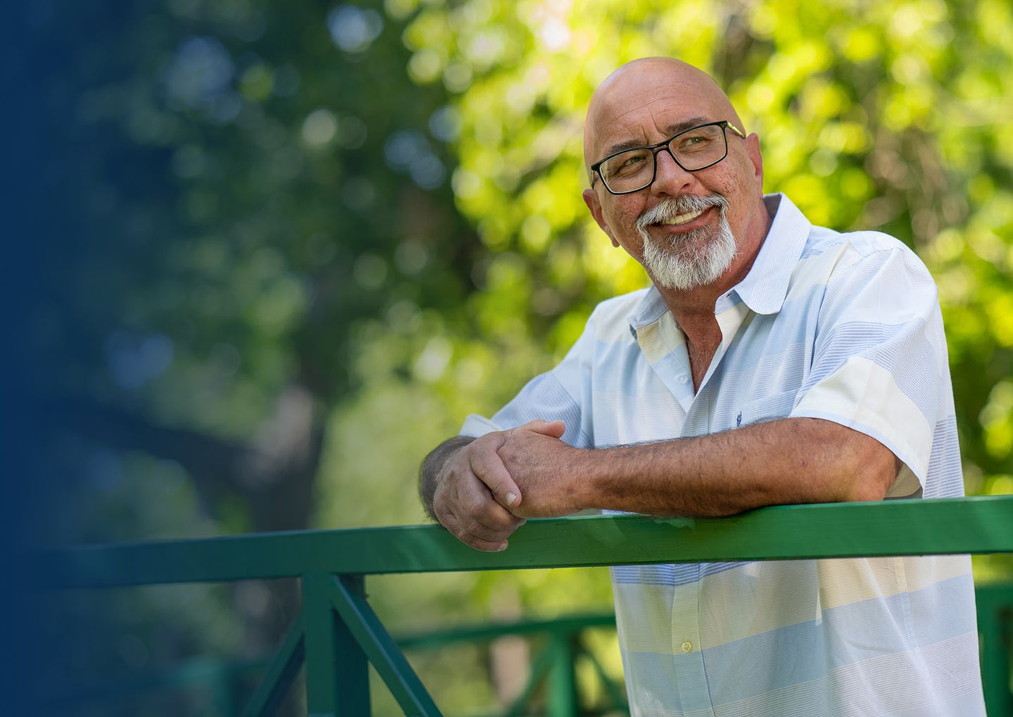 An older man in a short sleeve shirt is smiling outdoors while leaning on a green railing, representing confidence and positive outcomes after assessing dental implant eligibility.