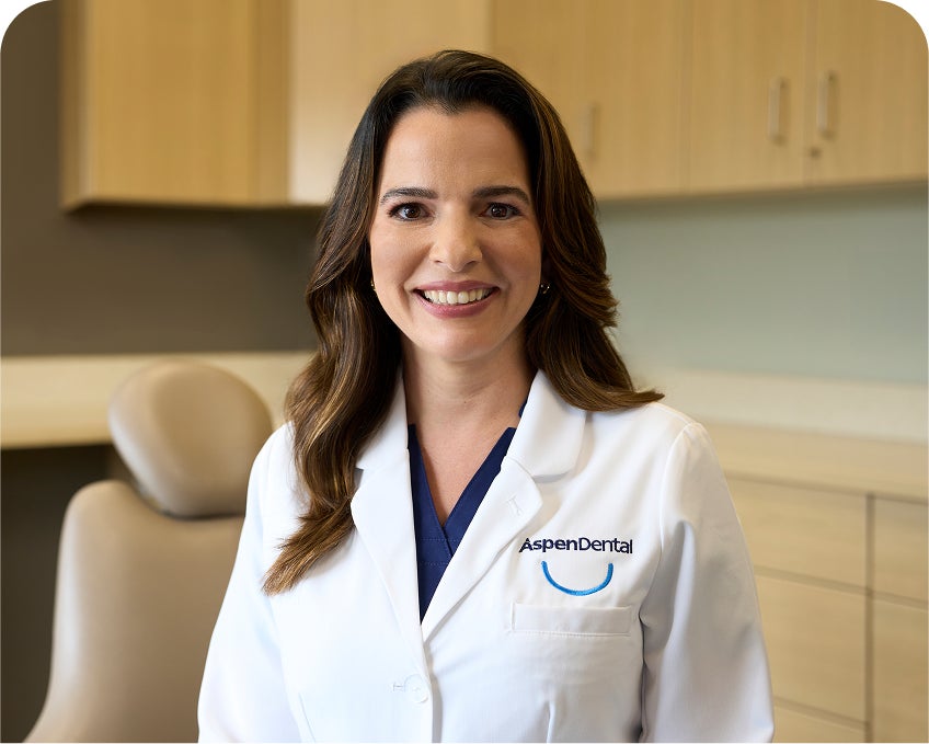 A smiling Aspen Dental dentist wearing a white coat stands in a modern dental exam room with cabinetry and a patient chair in the background.