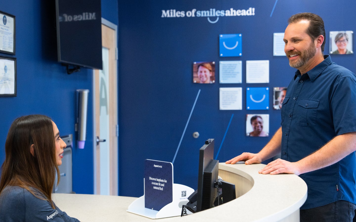 A patient smiles while speaking with an Aspen Dental team member at the front desk, with a blue wall behind them displaying the phrase “Miles of smiles ahead!”.