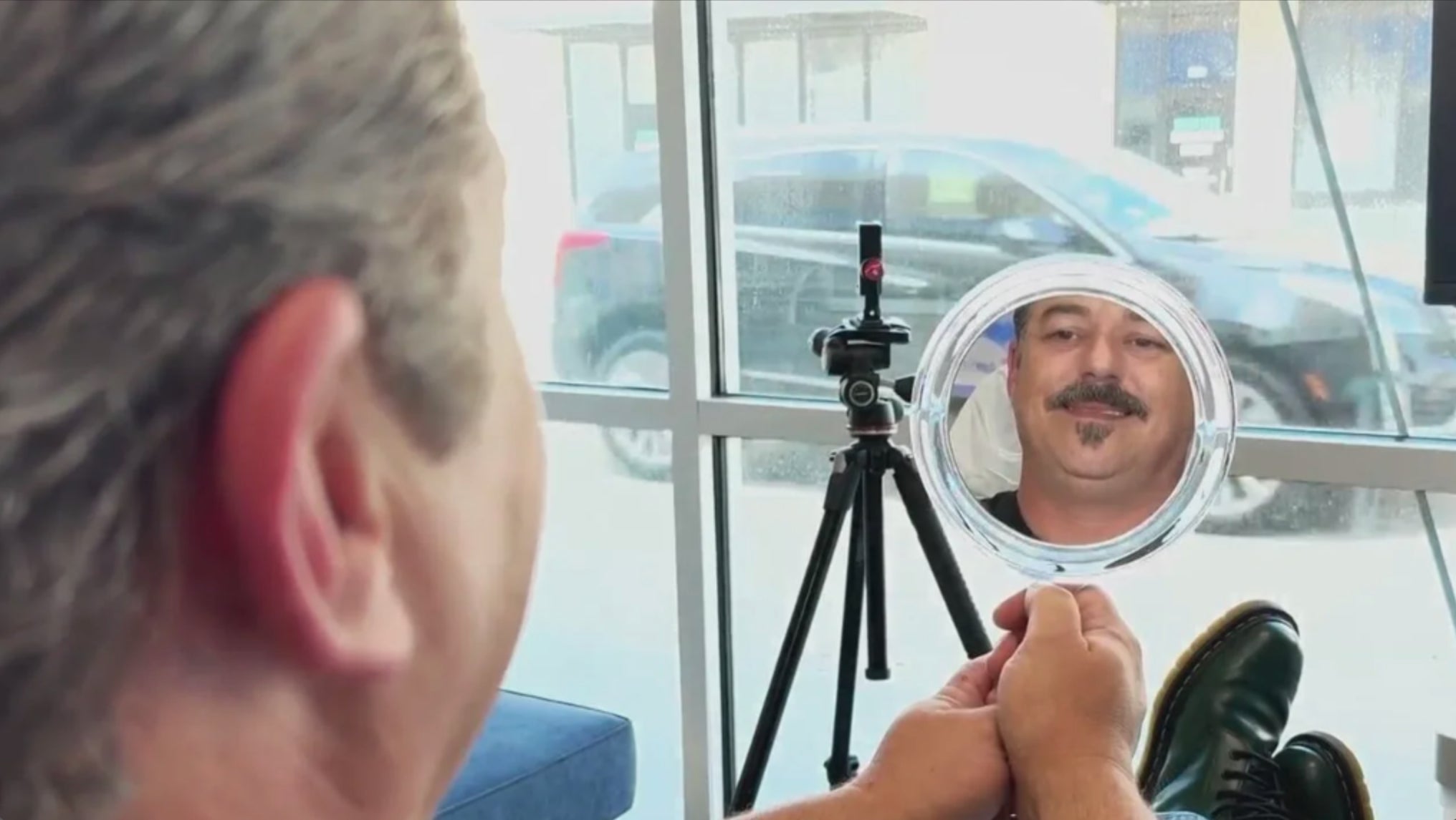A person holds a handheld mirror reflecting a smiling man with a mustache