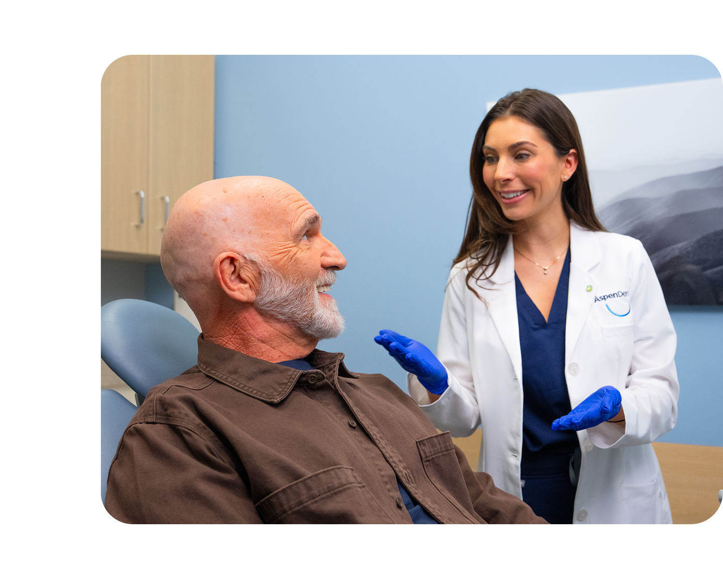 Aspen Dental dentist wearing gloves talks with a male patient who is smiling while seated in the dental chair.