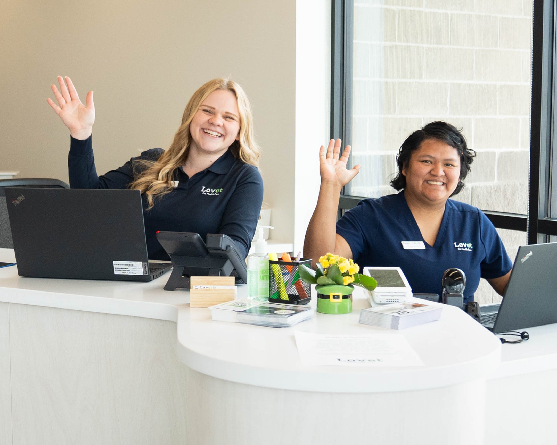 Two women waving while sitting at a desk with laptops.