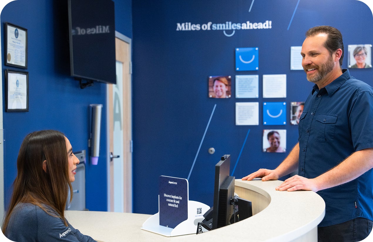 A smiling patient speaks with an Aspen Dental team member at the front desk, with a blue wall behind them displaying the phrase “Miles of smiles ahead!” and patient photos.