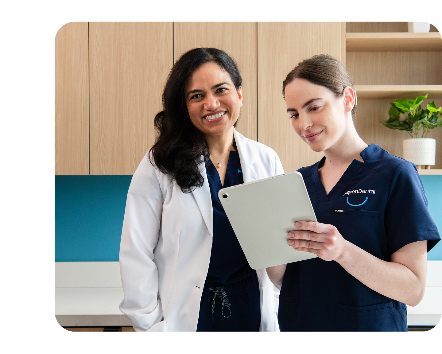 Two Aspen Dental team members, one in a white lab coat and one in scrubs, smiling while reviewing a patient chart on a digital tablet.