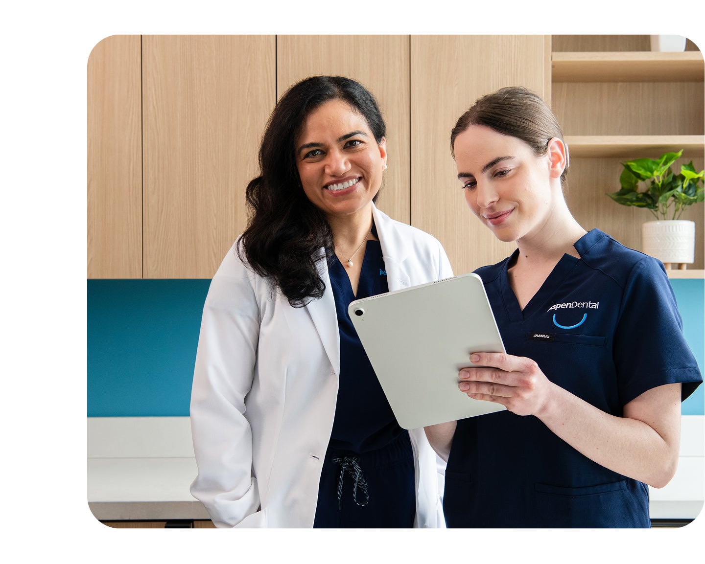 Two Aspen Dental team members, one in a white lab coat and one in scrubs, smiling while reviewing a patient chart on a digital tablet.