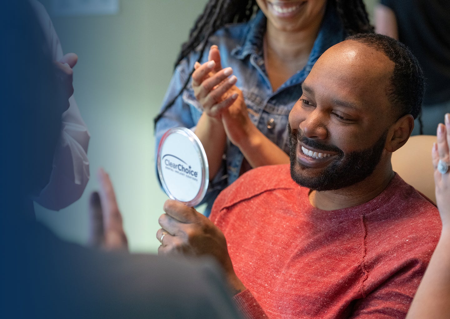 A male patient in a red shirt smiles into a hand held mirror as he celebrates his new dental implants with his prosthodontist and friends beside him.