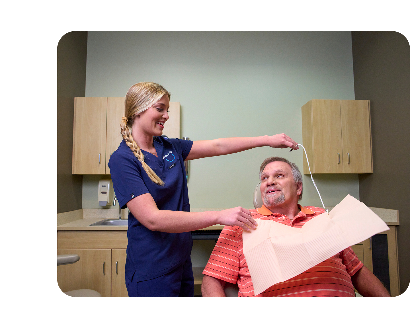 An Aspen Dental assistant in navy scrubs places a protective bib on a seated male patient in a dental exam room, preparing him for periodontal treatment.