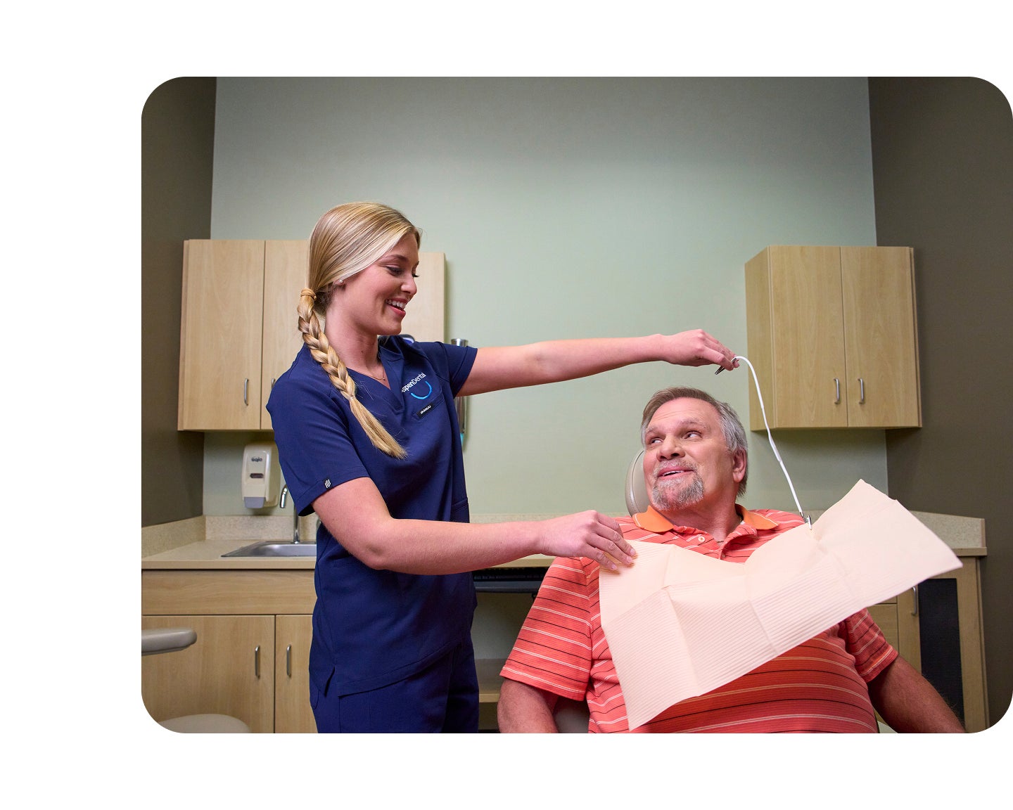 An Aspen Dental assistant in navy scrubs places a protective bib on a seated male patient in a dental exam room, preparing him for periodontal treatment.
