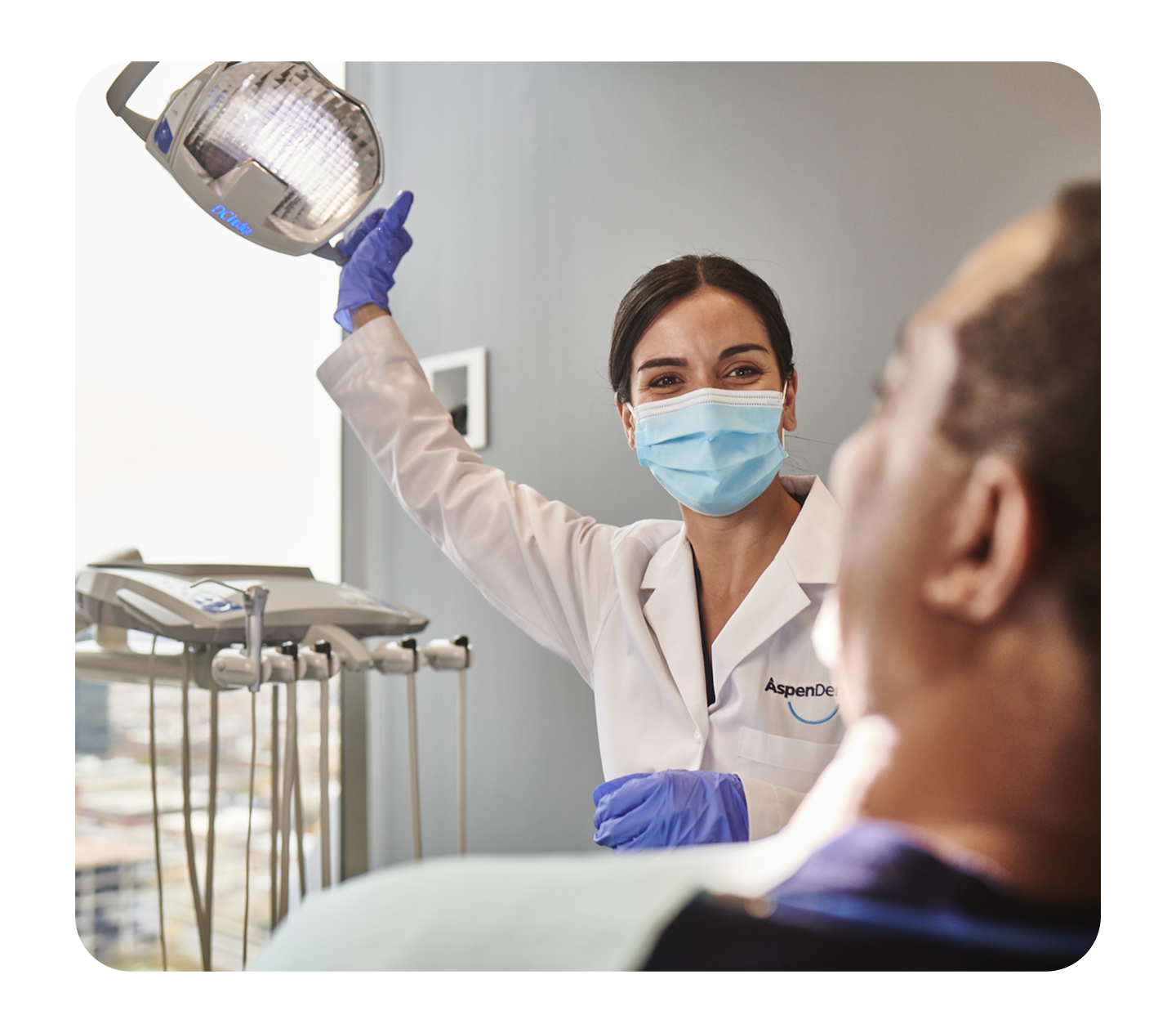 An Aspen Dental dentist wearing a mask and gloves adjusts an overhead exam light while smiling at a patient seated in the dental chair.