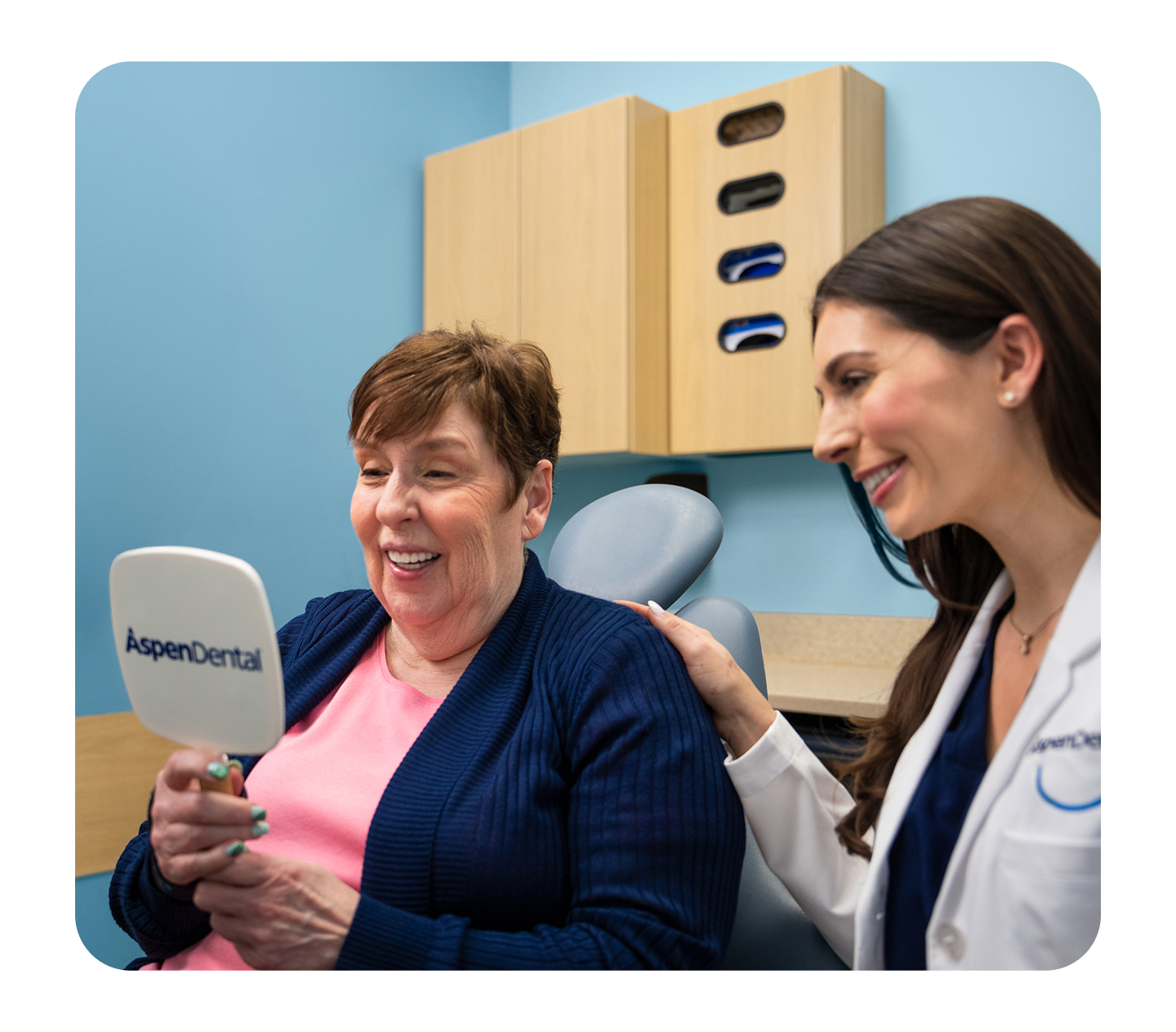 Two people in a dental office; one holds an Aspen Dental mirror while the other stands beside them with a hand on their shoulder.