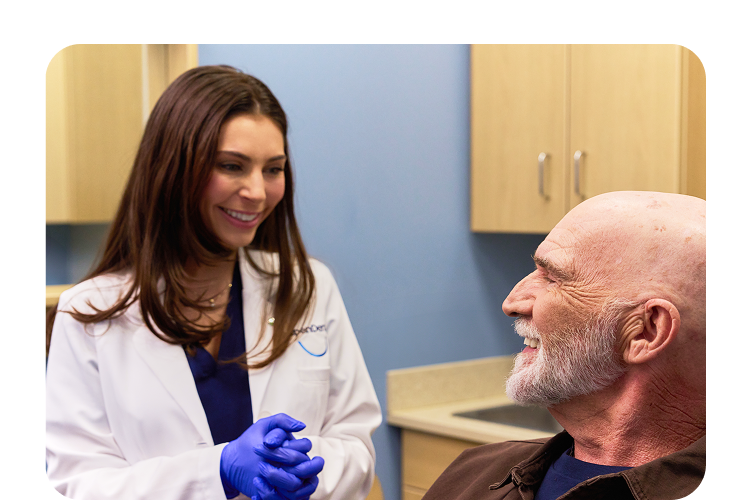 A female dentist in a white lab coat and blue gloves smiling and talking to a male patient with a beard, who is also smiling.