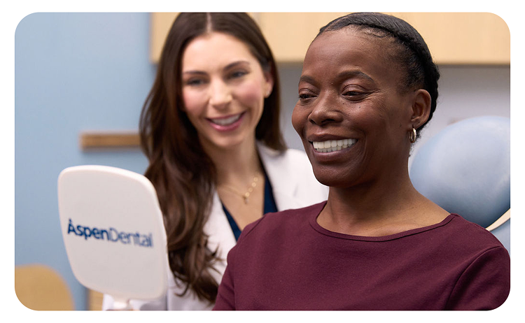 An Aspen Dental doctor in a white coat and blue gloves is smiling while having a discussion with a woman sitting in the dental chair.