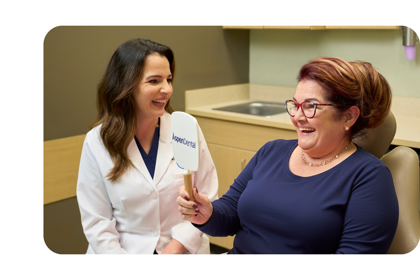 A smiling patient looks into an Aspen Dental mirror while a dentist in a white coat sits beside her, sharing a joyful moment during a cosmetic dentistry consultation.