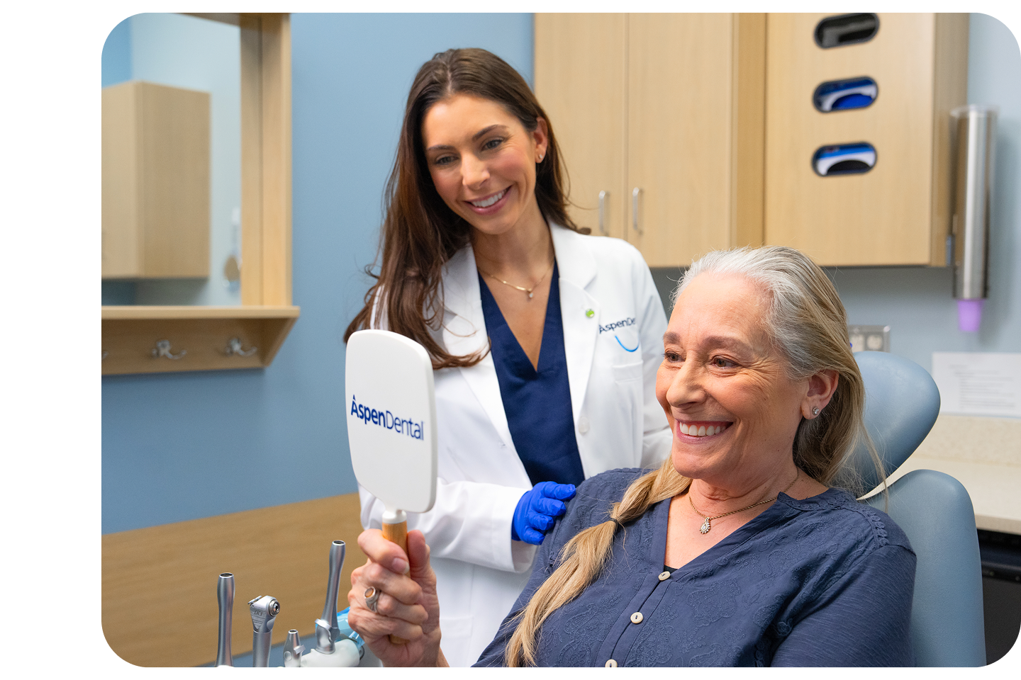 An older woman with long gray hair smiling brightly while looking at her reflection in a handheld mirror, while an Aspen Dental doctor stands smiling behind her.