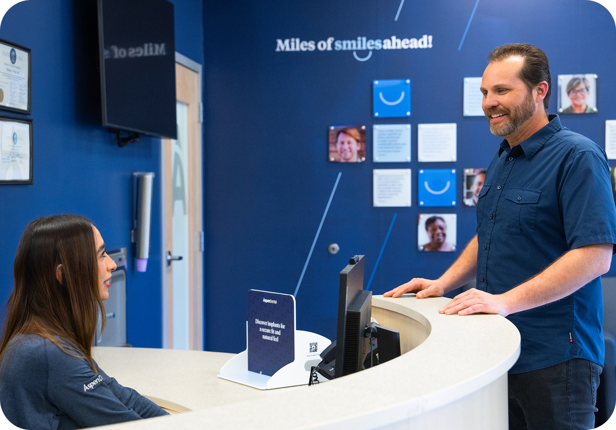 Male patient standing at the Aspen Dental front desk smiling and speaking with a receptionist in a blue office with “Miles of smiles ahead!” on the wall.