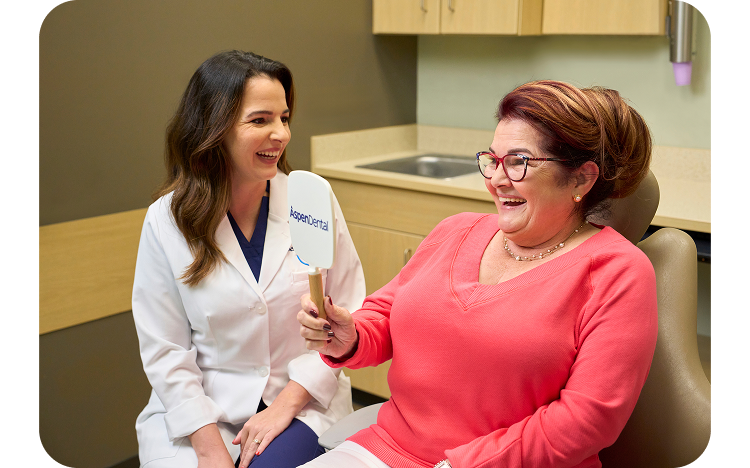 A happy patient wearing glasses smiles while looking into an Aspen Dental mirror, as a dentist in a white coat sits beside her and smiles during the appointment.