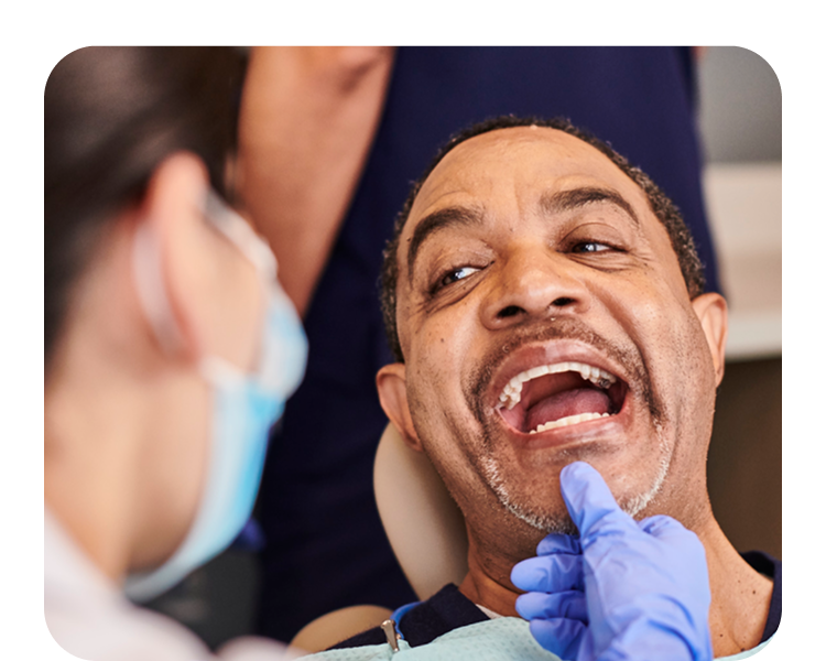 A male patient with a mustache is seated in a dental chair, smiling with his mouth open, as a dentist in a mask and blue glove points to a tooth on his lower jaw during an exam.