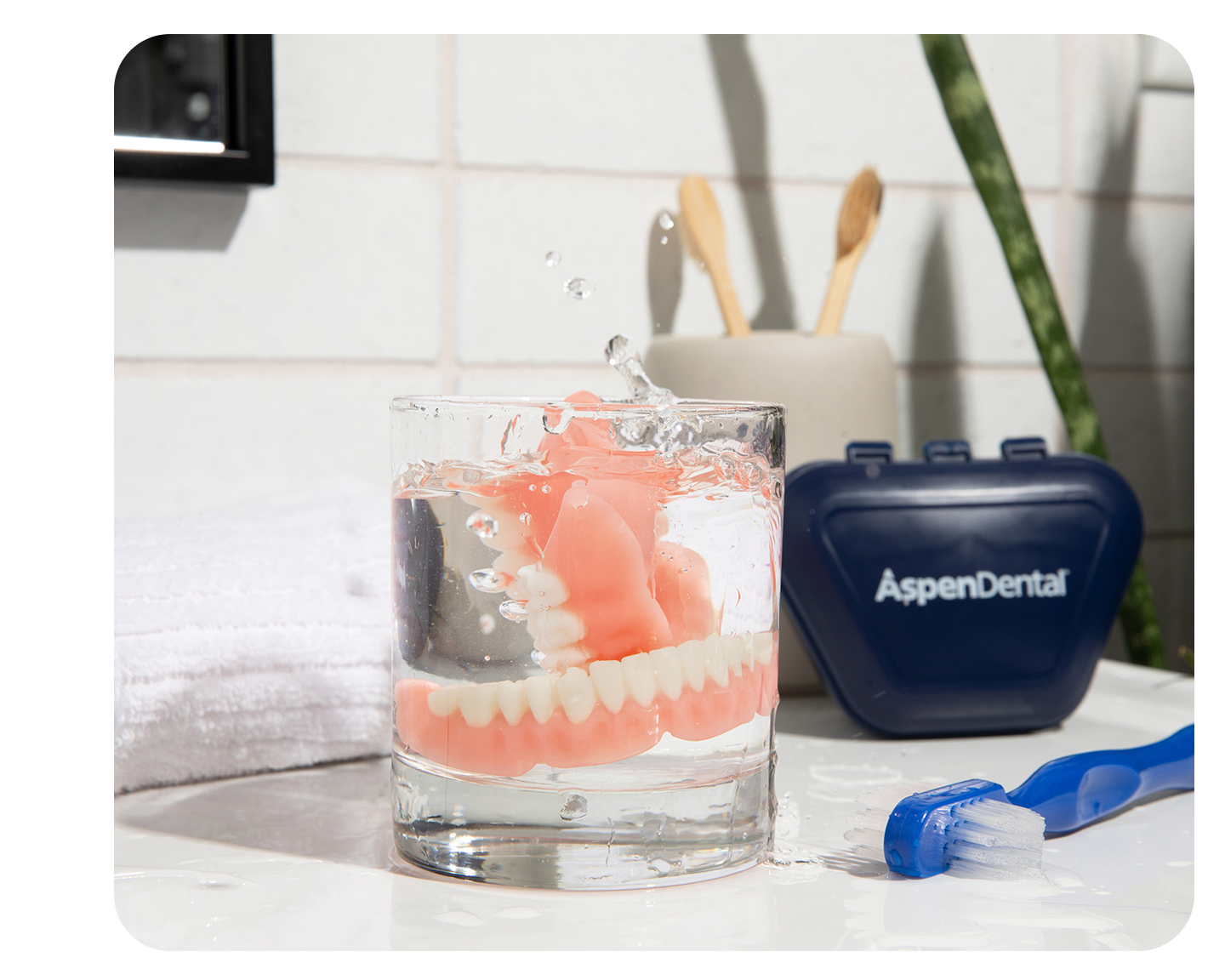 Dentures in a glass of water on a bathroom counter with a blue toothbrush, Aspen Dental container, wooden toothbrushes, and a white towel.