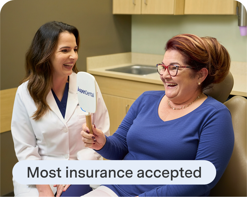 A smiling patient holds an Aspen Dental mirror while sitting with a dentist in a dental exam room, with text reading “Most insurance accepted” displayed below.