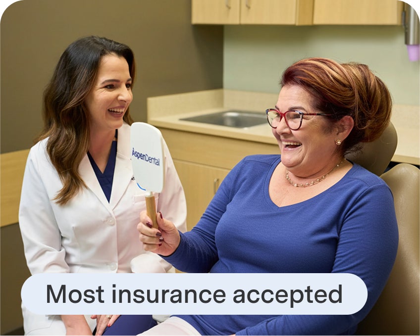 A smiling patient holds an Aspen Dental mirror while sitting with a dentist in a dental exam room, with text reading “Most insurance accepted” displayed below.
