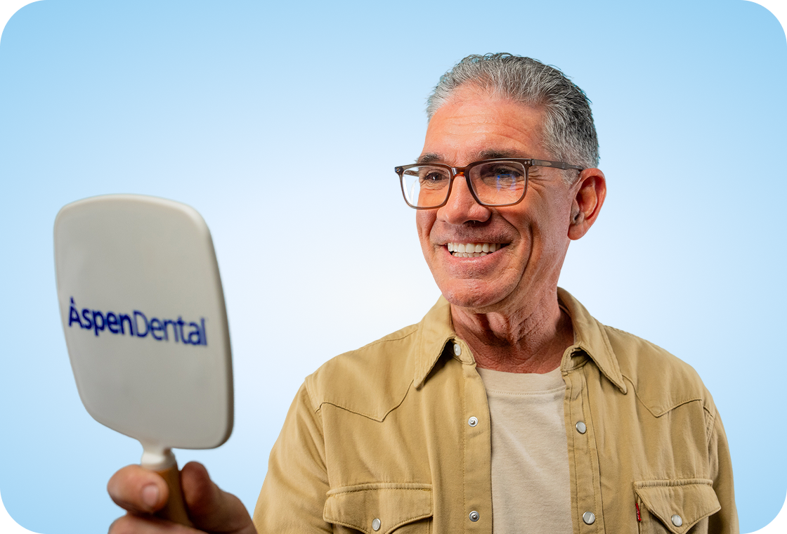 An older person with gray hair and glasses is holding a hand mirror with the Aspen Dental logo on it, looking at their reflection.