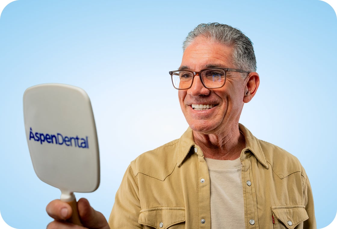 An older person with gray hair and glasses is holding a hand mirror with the Aspen Dental logo on it, looking at their reflection.