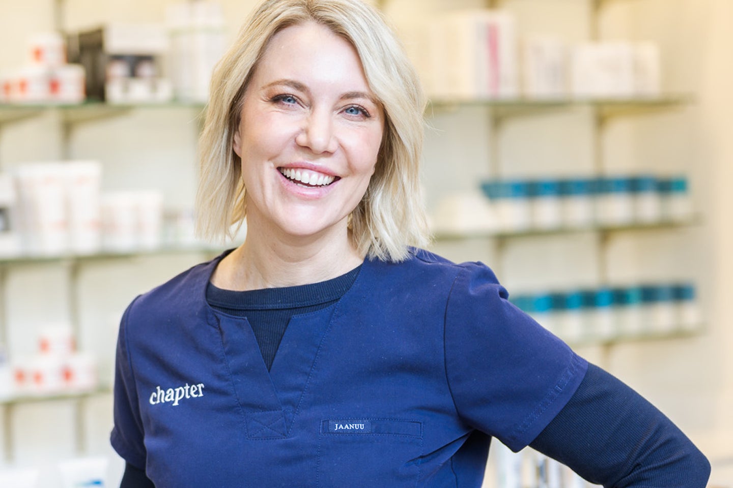 A smiling woman with short blonde hair smiling and wearing blue scrubs in front of a shelf of skincare products