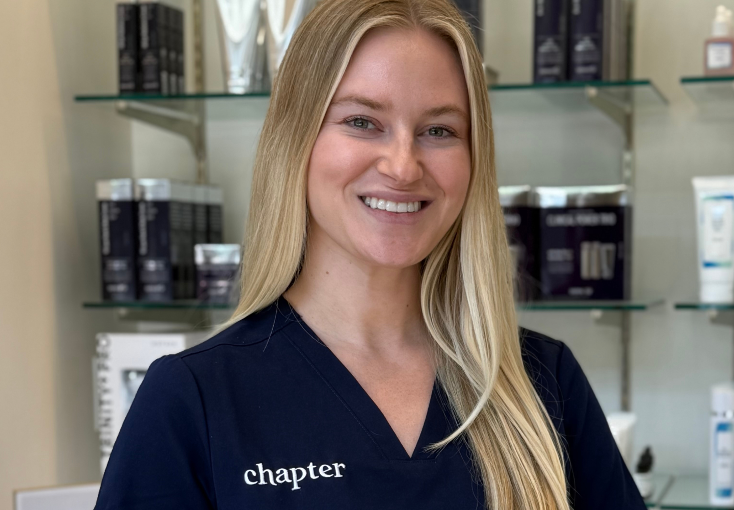 A woman in a navy blue chapter scrub with long blonde hair is smiling for the camera