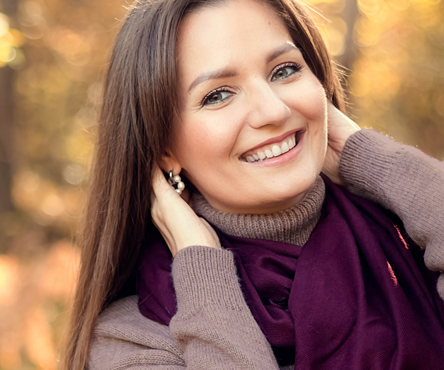 A woman with brown hair, wearing a sweater and burgundy scarf, smiles against an autumnal forest background