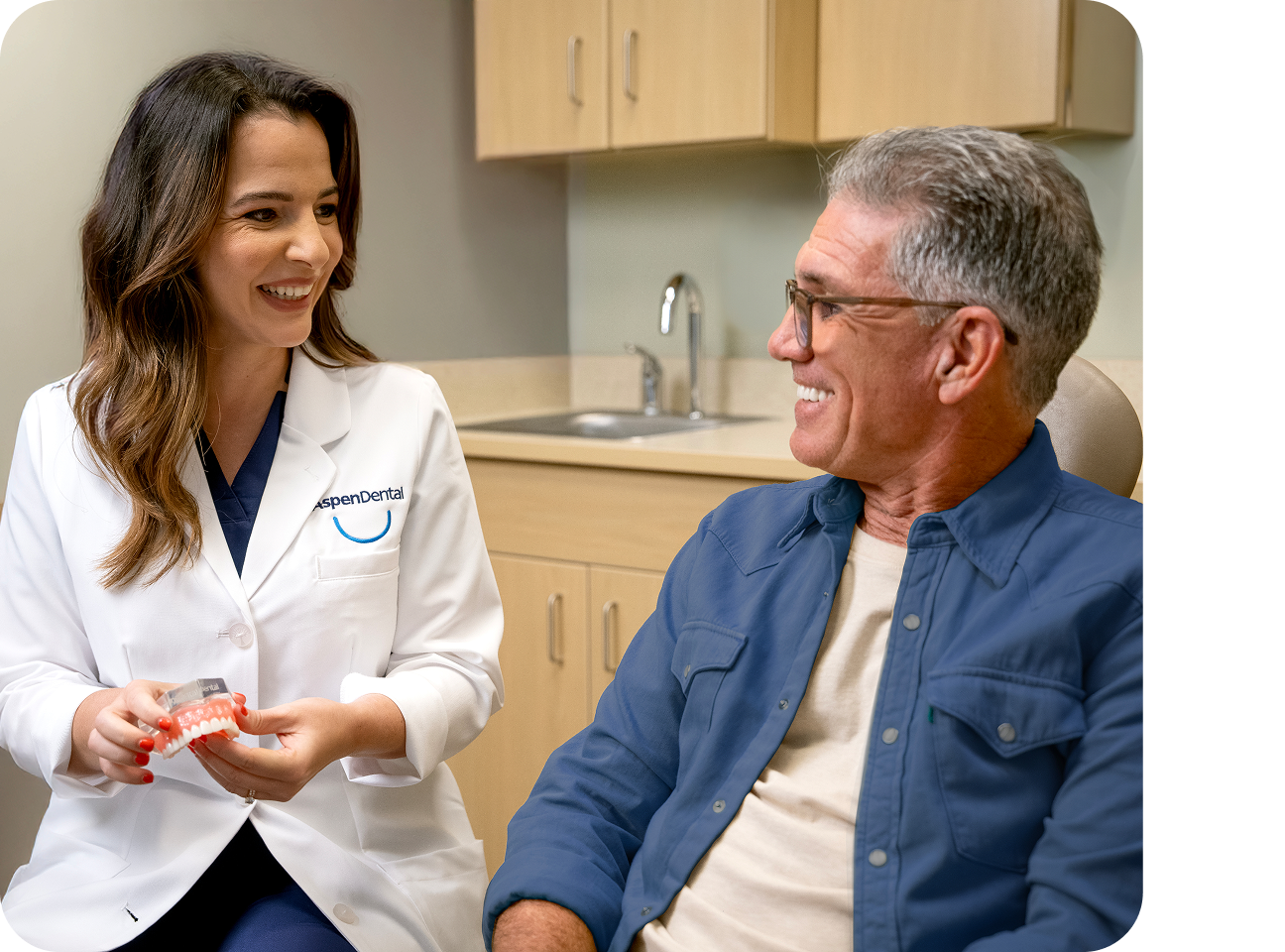 An Aspen Dental dentist smiles while holding a dental model and explaining treatment options to a male patient seated in a dental exam chair.