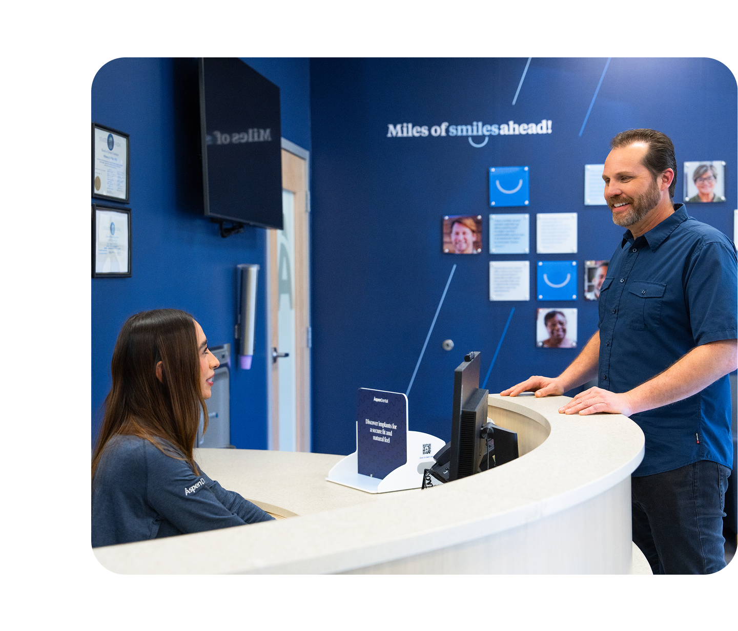 A smiling patient stands at the Aspen Dental front desk speaking with a team member, with a blue wall behind them displaying the phrase “Miles of smiles ahead!”